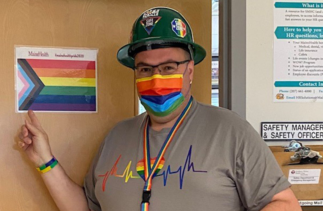 a man wearing a pride-themed facemask, pride-themed t-shirt while pointing to a pride flag hanging on a door