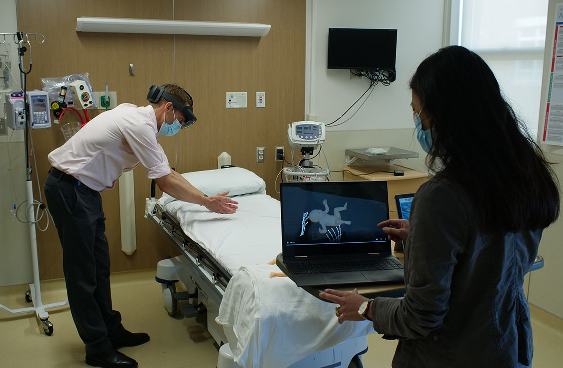a man wearing VR goggles next to a gurney while a woman views a computer simulation of an infant on a laptop