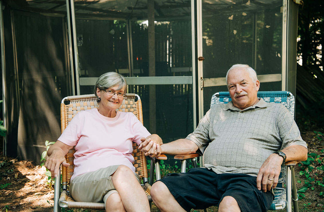 older man and woman sitting together and holding hands outside in a wooded area
