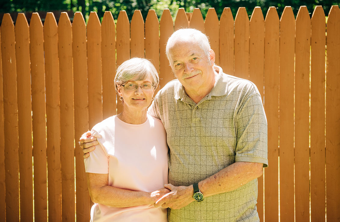 older husband and wife standing together in front of tall wooden fence on a sunny day