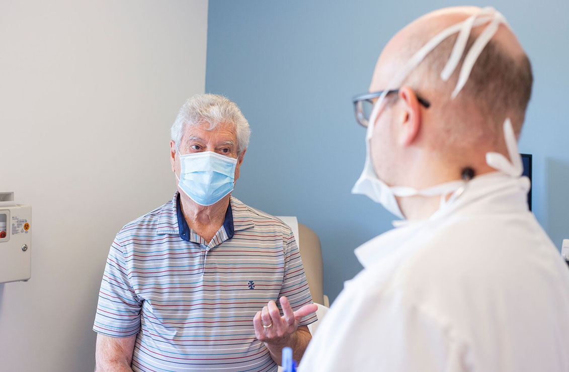 older patient wearing a mask and speaking to a doctor in an exam room