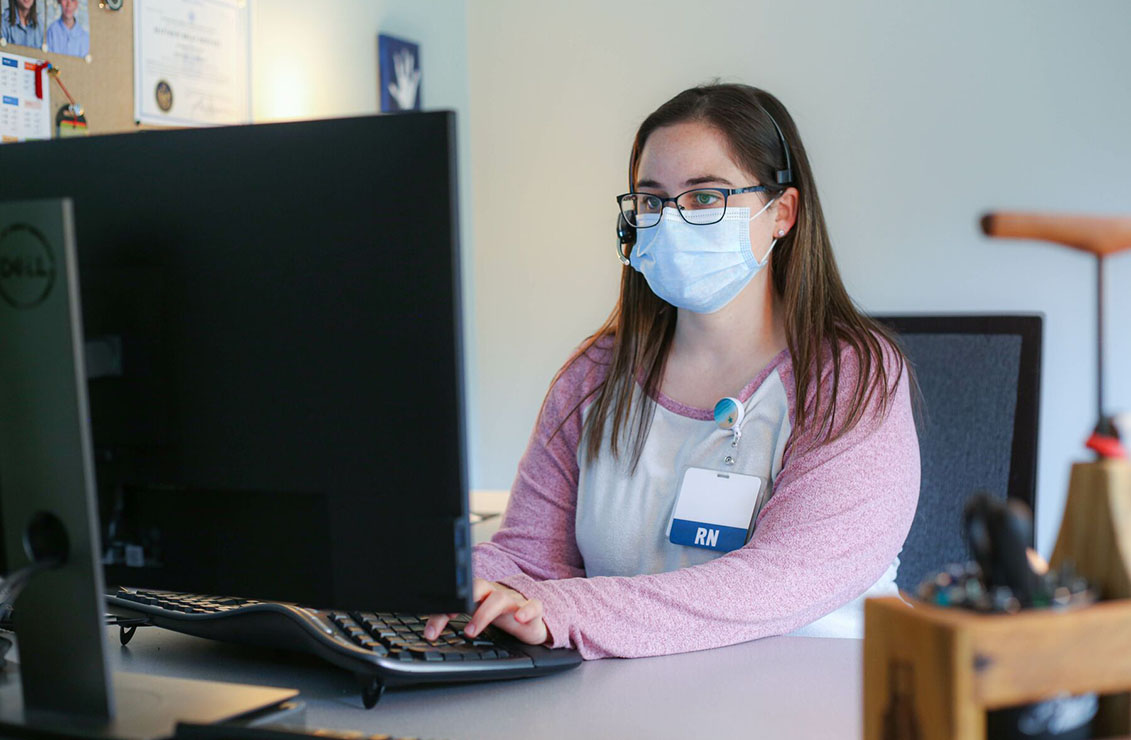 nurse wearing a mask, sitting and typing on a keyboard while looking at a computer screen