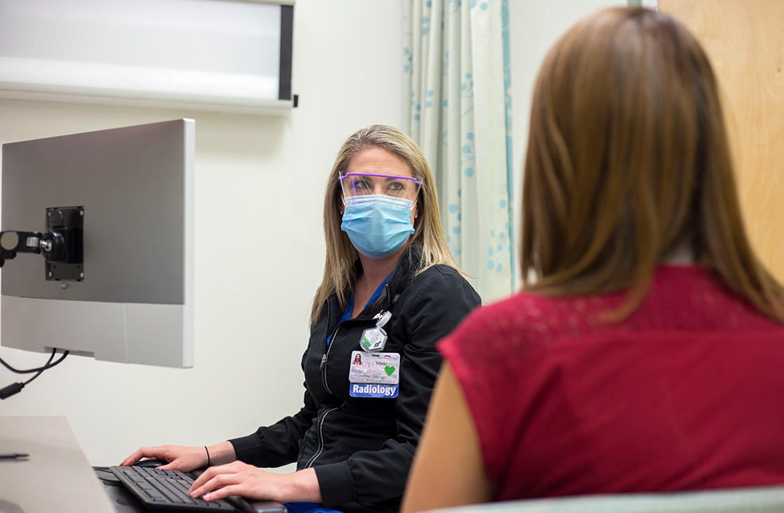 woman wearing a surgical mask and sitting at a computer workstation next to a patient