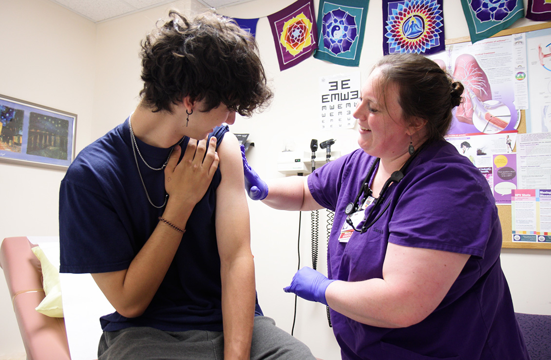 A woman care team member examines a teen, male student in high-school exam room.