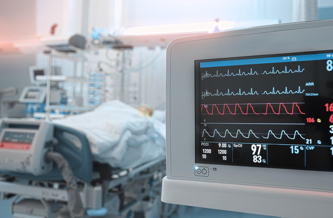 close-up view of a cardiac monitor with a patient lying in a hospital bed behind it